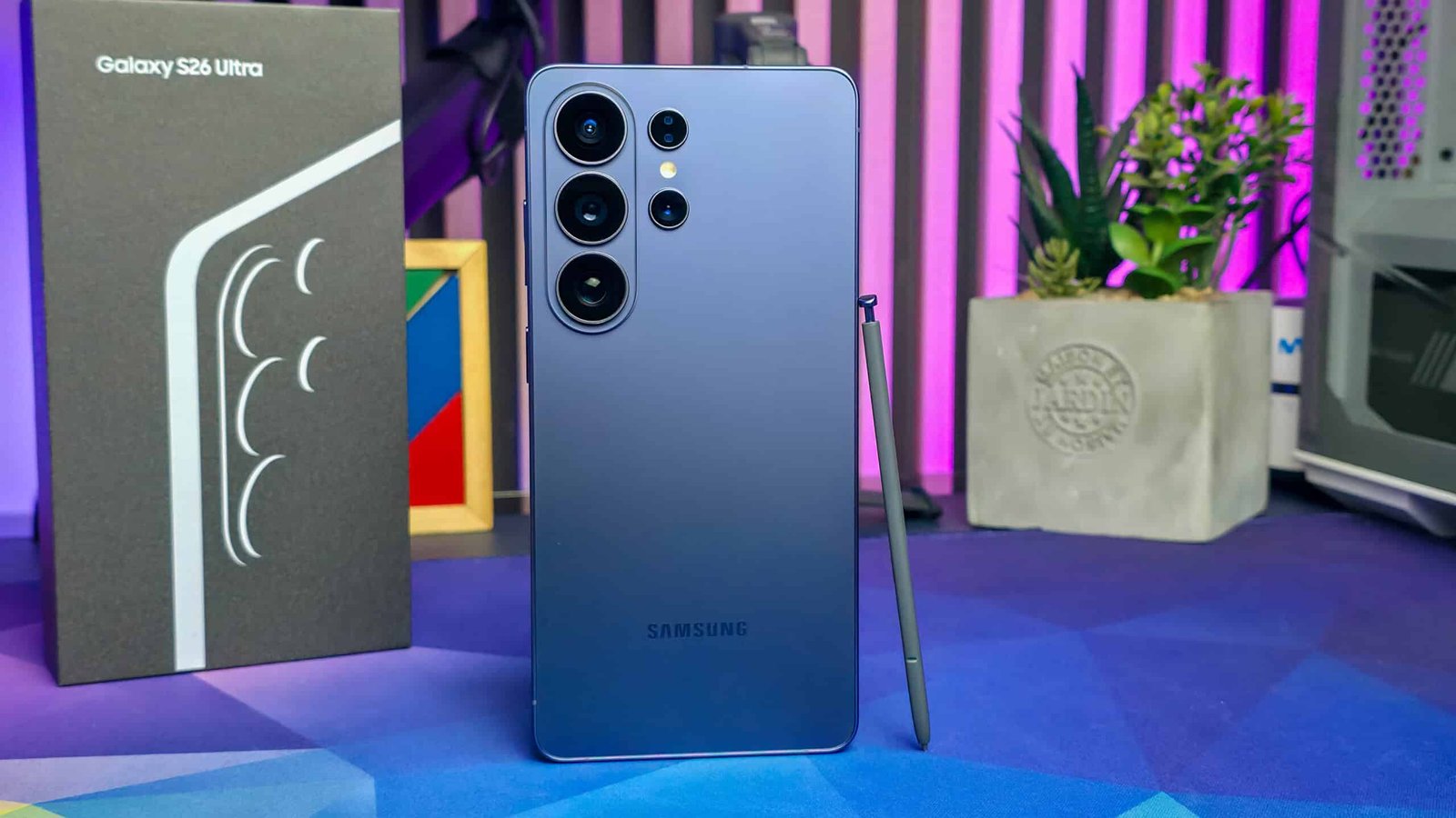 Vista trasera del Samsung Galaxy S26 Ultra mostrando su módulo de cámaras múltiples, acompañado del S Pen y su caja, en un entorno de escritorio con luces LED y decoración